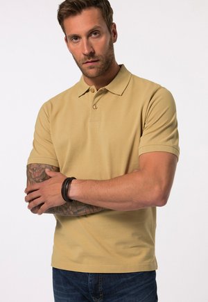Beige polo shirt with short sleeves, two-button placket, and textured fabric. Model has tattoos on arms and wears a dark bracelet.