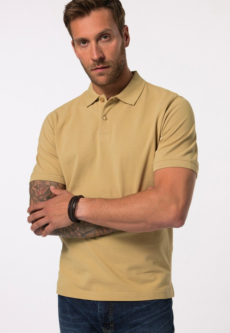 Béžové polo tričko s krátkými rukávy, dvoubarevnou knoflíkovou légou a texturovanou tkaninou. Model má tetování na rukou a nosí tmavý náramek.