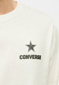 Λευκό μπλουζάκι με στρογγυλή λαιμόκοψη, με ένα μαύρο αστέρι που παρουσιάζει ένα τοπογραφικό μοτίβο πάνω από τη λέξη "CONVERSE" τυπωμένη στο στήθος.