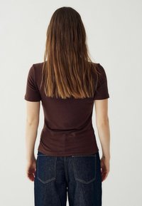 Rjava kratka majica s kratkimi rokavi, izdelana iz mehkega materiala, prilega se telesu, vidni šivi, v kombinaciji z visokimi temnimi džins hlačami.