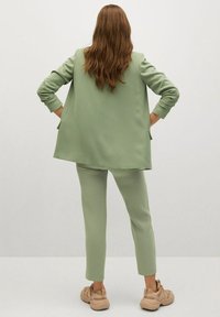 Costume vert avec une veste surdimensionnée et un pantalon slim, fabriqué en tissu lisse. Associé à des baskets beiges. Vue de dos, fond uni.