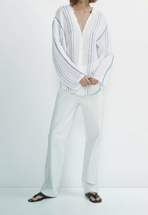 Kobieta ubrana w luźną białą koszulę na guziki w niebieskie paski, szerokie białe spodnie oraz czarne sandały z odkrytymi palcami, stojąca na białym tle.