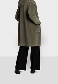 Cappotto in tessuto verde oliva, morbido e peloso, con cappuccio, maniche lunghe e tasche laterali. Abbinato a pantaloni neri a gamba larga e stivali neri chunky.