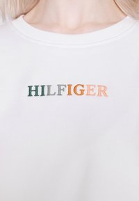 Sudadera blanca con cuello acanalado que presenta "HILFIGER" bordado en letras verdes, naranjas y beige, sobre una tela lisa y texturizada.