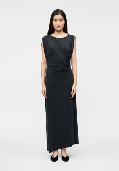 Femme aux longs cheveux noirs portant une robe maxi noire sans manches avec un détail froncé sur le côté, debout droite sur un fond blanc.