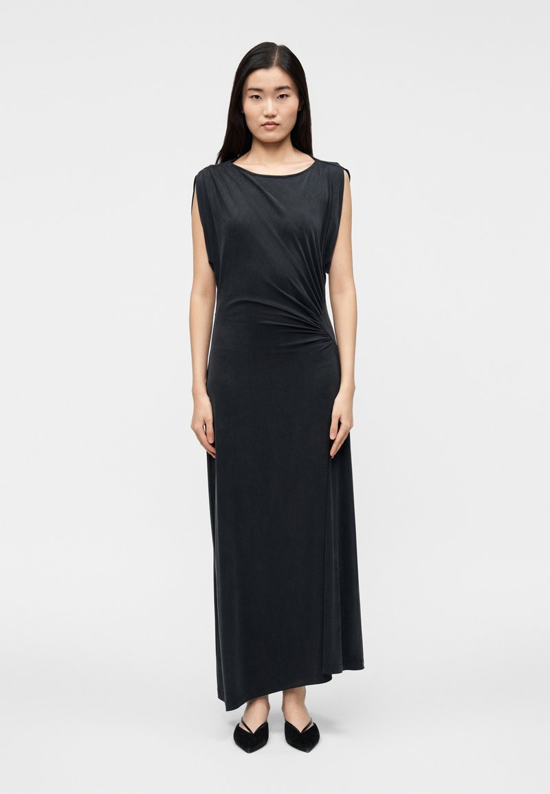 Woman with long dark hair wearing a sleeveless black maxi dress with ruched side detail, standing straight on white background.