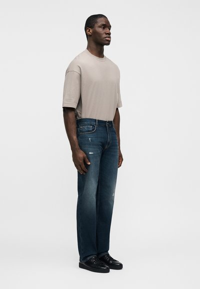 Άνδρας που φοράει μπεζ υπερμεγέθη μπλουζάκι, σκούρο μπλε τζιν παντελόνι ίσιας γραμμής με ελαφριά φθορά και μαύρα αθλητικά παπούτσια χαμηλής κορυφής, στέκεται κοιτάζοντας δεξιά.