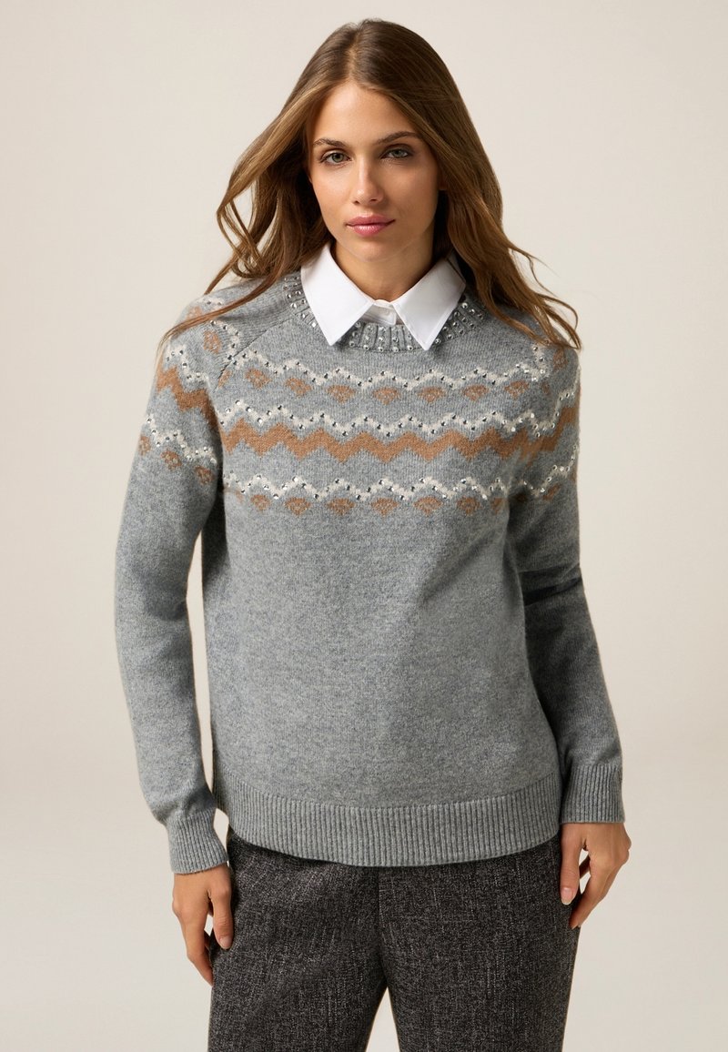 Suéter de punto gris con un patrón geométrico en beige y blanco, con cuello redondo y dobladillo y puños acanalados, superpuesto a una camisa blanca con cuello.