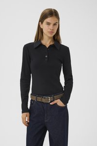 Woman with straight hair wearing a black long-sleeve collared shirt, blue jeans, and a brown belt, standing with one hand in pocket.