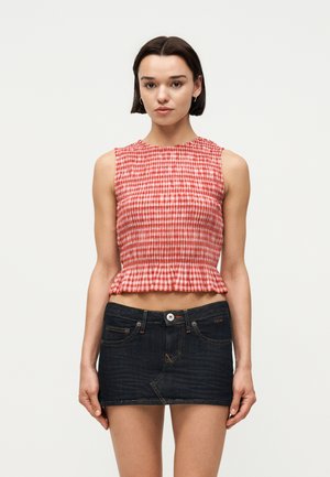 Kobieta o krótkich ciemnych włosach, ubrana w czerwoną i białą kraciastą bluzkę bez rękawów oraz ciemną mini spódniczkę z denimu, stojąca na jednolitym tle.