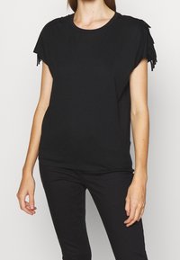 Svart kortärmad t-shirt med fransade detaljer. Mjuk kvalitet, avslappnad passform, rund halsringning. Kombinerad med svarta byxor för en monokromatisk look.