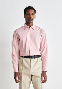 Camicia rosa chiaro a maniche lunghe con colletto classico, con un piccolo logo ricamato, indossata con pantaloni leggeri a fantasia e una cintura nera.