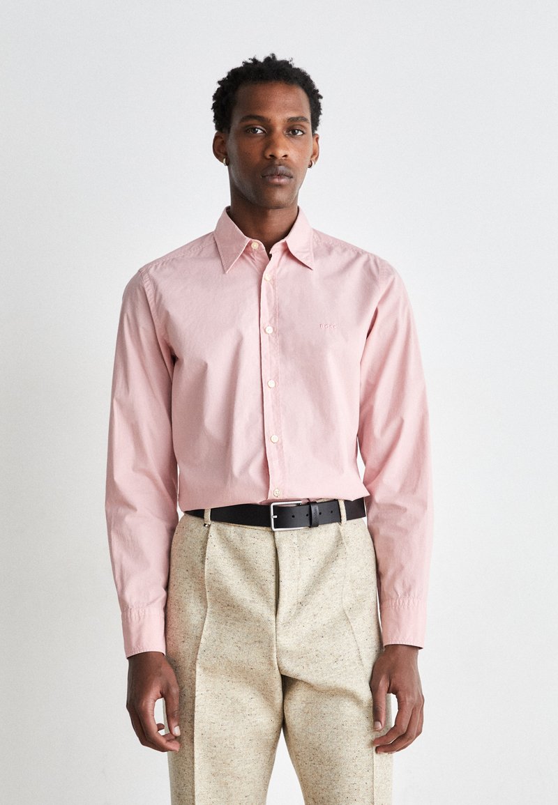 Camicia rosa chiaro a maniche lunghe con colletto classico, con un piccolo logo ricamato, indossata con pantaloni leggeri a fantasia e una cintura nera.