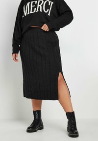 Mustat neulotut villapaidat, jossa on "MERCI"-teksti, yhdistetty mustaan perhosneuleiseen midi-hameeseen, jossa on sivuaukko, ja käytetty mustien nauhakenkien kanssa.