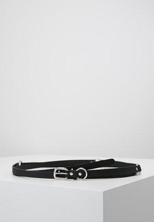 Ceinture fine en cuir noir avec boucle argentée et trous ajustables, placée enroulée sur une surface blanche avec un fond gris.