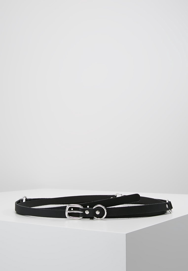 Ceinture fine en cuir noir avec boucle argentée et trous ajustables, placée enroulée sur une surface blanche avec un fond gris.