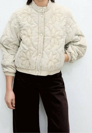 Personne portant une veste matelassée beige avec broderie florale et un pantalon large foncé, debout devant un fond blanc uni.