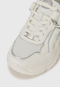 White sneaker featuring a textured upper, shiny silver accents, and round laces. Branding tag is visible on the side. Thick sole for support.