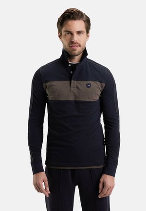 Un homme portant un polo à manches longues sombre avec une bande marron sur la poitrine et un col montant, se tenant devant un fond uni.