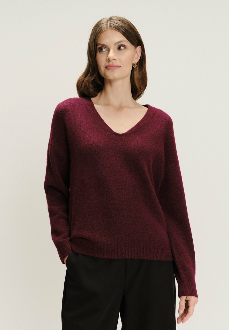 Vrouw in een bordeaux V-hals trui met lange mouwen en zwarte broek, staande met één hand in de zak tegen een effen achtergrond.