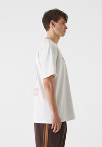 Biela bavlnená tričko s kratkým rukávom, na zadnej strane s ružovým grafickým textom. Jednoduchý dizajn s voľným strihom a minimálnou textúrou.