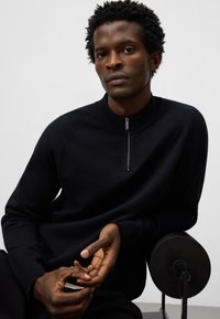 Svart stickad halvdraget tröja med ribbat krage och raglanärmar, tillsammans med mörka byxor, bärs av en person med kort, texturerat hår.