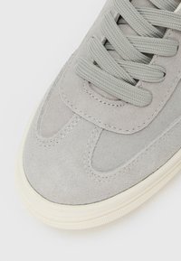 Close-up of light grey suede sneaker with thick white sole and matching grey flat woven laces on white background.