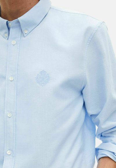 Camisa de botón azul claro con cuello abotonado, emblema bordado en el pecho y mangas remangadas en el brazo derecho.