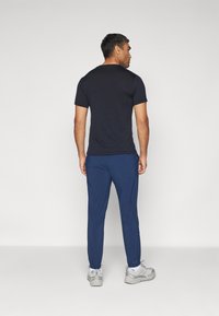 Chemise noire à manches courtes pour homme, associée à un pantalon de sport bleu et des baskets grises, vue de dos, mettant en avant une coupe décontractée.