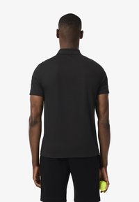 Polo shirt nero a maniche corte con colletto classico, texture liscia e design aderente. Indossato con pantaloni corti neri, mentre tiene una pallina da tennis.