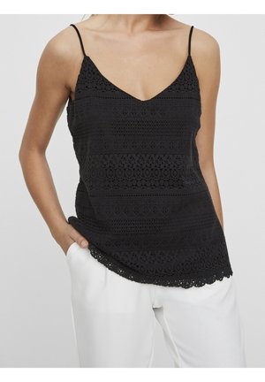 Top negro sin mangas con tirantes ajustables, que presenta un intrincado patrón de encaje y un dobladillo con forma de ondas, usado sobre pantalones blancos.