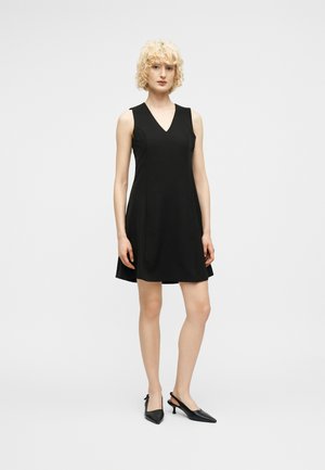 Kvinde med krøllet blondt hår iført en ærmeløs sort V-hals kjole og sorte slingback-hæle stående mod en lys baggrund.