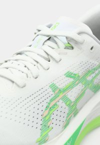 White athletic shoe with a mesh upper, green accents, and textured patterns. Features flat laces and a lightweight, cushioned sole.