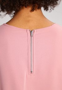 Rosa plagg med en mjuk textur som har en central dragkedja bak. Kragen har en rundad halsringning.