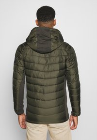 Veste matelassée vert olive avec capuche, affichant un design en quilté et des panneaux latéraux gris contrastants. Texture douce et poignets élastiques.