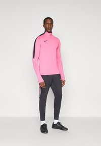 Ροδοκόκκινη αθλητική μπλούζα με μακρύ μανίκι και ψηλό γιακά, συνδυασμένη με σκούρες γκρίζες φόρμες που διαθέτουν ροζ λεπτομέρειες. Μαύρα αθλητικά παπούτσια.