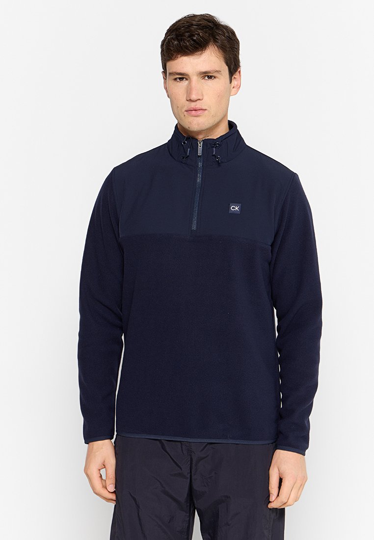 Marineblauwe fleece pullover met halve rits en een gladde textuur, voorzien van een gekleurde bovenpan en een CK-logo op de borst.
