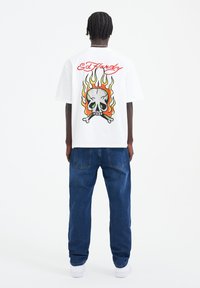 White cotton t-shirt featuring a large graphic of a skull with flames in green, orange, and yellow on the back. Relaxed fit, short sleeves.