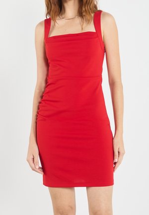 Kobieta w przylegającej, bezrękawowej sukience o kwadratowym dekolcie w jasnoczerwonym kolorze stojąca na tle jednolitego, jasnego tła.