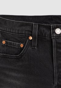 Gros plan sur un jean en denim noir montrant la ceinture, la passants de ceinture, la poche avant avec des rivets en cuivre et une fermeture à bouton argentée.