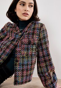 Giacca in tweed multicolore caratterizzata da un tessuto testurizzato con un motivo intrecciato, scollatura rotonda e bottoni neri, indossata con un dolcevita scuro.