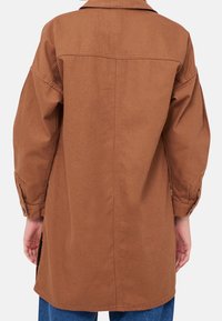 Osoba oblečená v hnědé kožené bundě s knoflíky a dlouhým rukávem, s límcem, viděná zezadu, oblečená do modrých džínů na bílém pozadí.