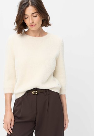 Vrouw met bruin haar die een wit fuzzy trui met driekwart mouwen en donkerbruine broek met een riem draagt, kijkt naar beneden.