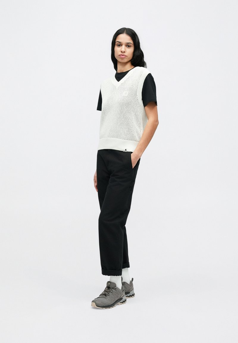 Gilet bianco in rete indossato sopra una maglietta nera a maniche corte, abbinato a pantaloni neri e scarpe sportive grigie. Caratterizzato da look e texture sovrapposti.