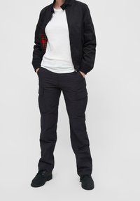 Schwarze Bomberjacke mit kariertem Innenfutter, weißem T-Shirt und schwarzen Cargo-Hosen mit mehreren Taschen. Stiefel in einer ähnlichen dunklen Farbe.