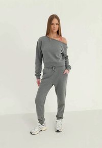 Jovem mulher em pé, vestindo um sweatshirt cinza sem ombros, calças de moletom combinando, um top desportivo rosa e tênis brancos, em um fundo claro.
