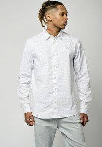 Chemise blanche à boutons avec un motif bleu moucheté, manches longues, col pointu et logo discret sur la poitrine. Associée à un pantalon gris clair.