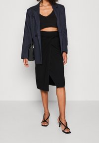 Haut court noir, jupe noire taille haute avec détail torsadé, et blazer bleu foncé. Assorti à des talons noirs à brides et un petit sac.