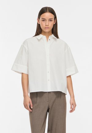 Femme aux cheveux bruns et raides porte une chemise blanche ample à manches courtes avec boutons et un pantalon marron sur mesure devant un fond blanc uni.
