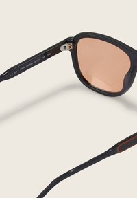 Czarne okulary przeciwsłoneczne z różowymi soczewkami, prostokątny kształt i kontrastowe pomarańczowe akcenty na zausznikach. Metalowe detale przy zawiasach.